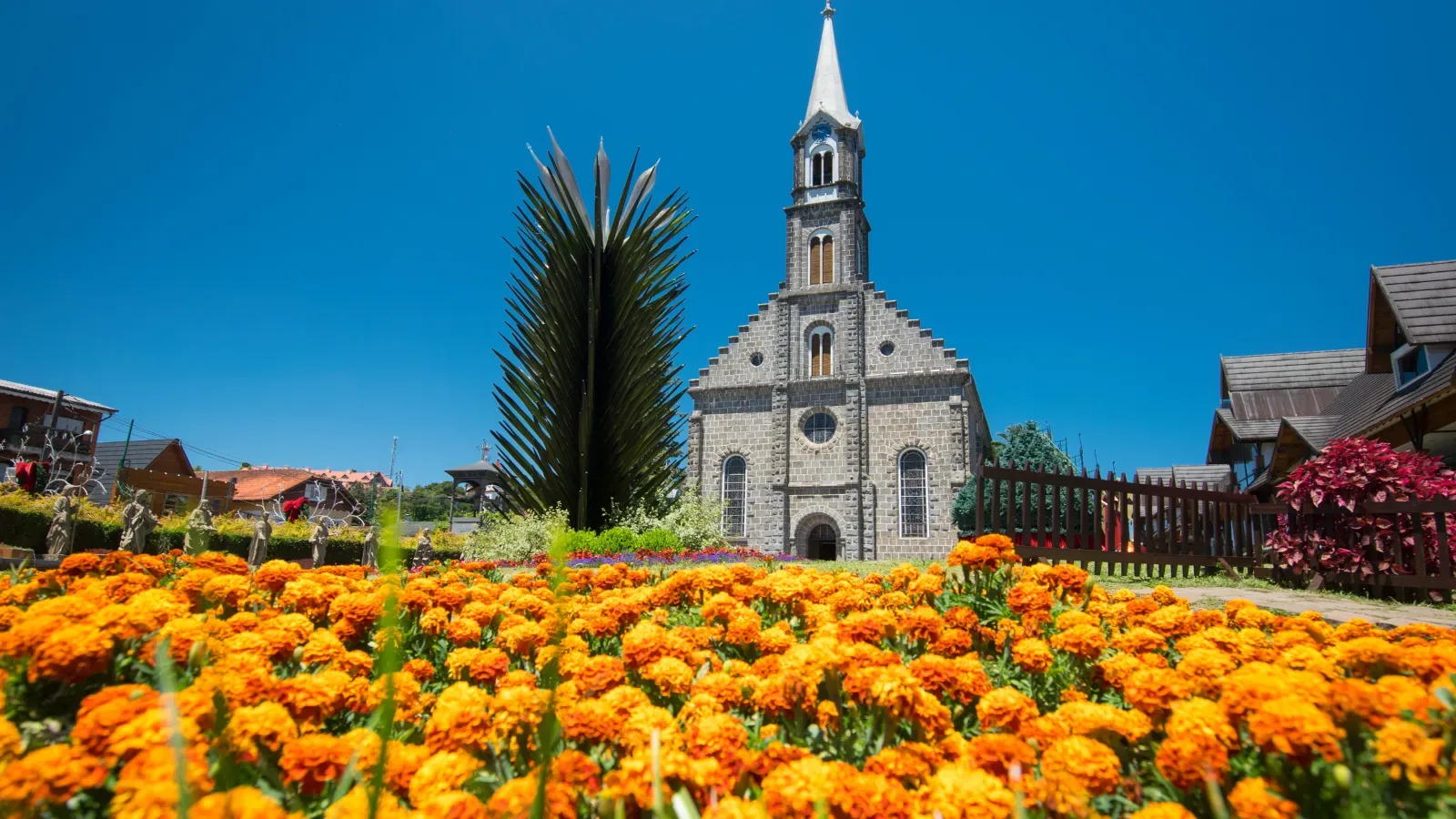 Imagem da igreja gótica de São Pedro em Gramado, com um céu azul e flores amarelas e laranjas na frente