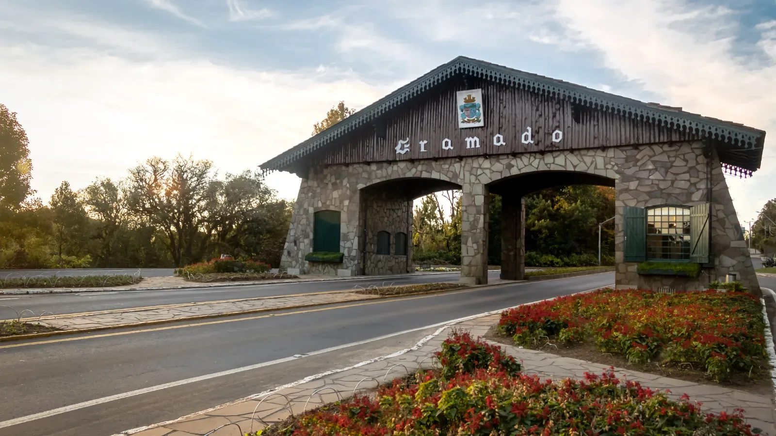 portico da cidade de gramado, feito de pedra em um dia ensolarado, a direita flores vermelhas do jardim