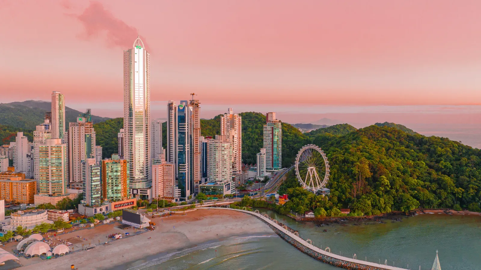 Vista de cima da cidade de Balneário Camboriú em um por do sol com tons rosas, mostrandos os prediso e a mata atlantica